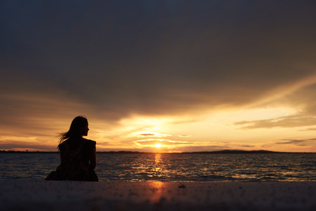 Back view silhouette of tourist woman sitting alone on seashore at water edge, enjoying beautiful view of sunset on dark evening sky background. Tourism and vacations concept.の写真素材