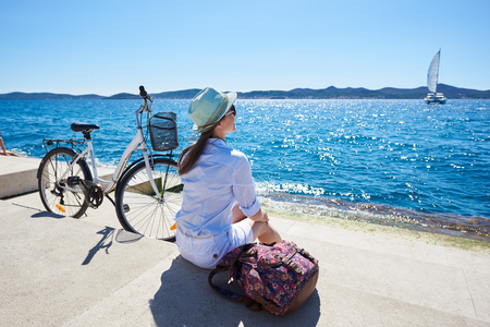 Perfect summer landscape on bright sunny day. Pretty smiling tourist girl in sunglasses with backpack sitting at bicycle on paved sidewalk under clear blue sky watching sailing ship in azure water.の写真素材