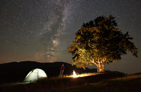 Happy female hiker relaxing at summer night camping in the mountains beside campfire, glowing tourist tent and big old tree. Back view of woman enjoying view of night sky full of stars and Milky way.の写真素材