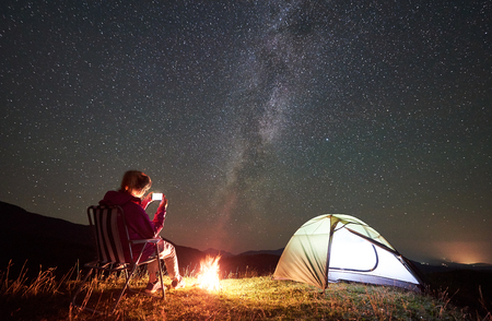 Female hiker having a rest at summer night camping in the mountains beside campfire and glowing tourist tent. Young woman sitting on chair, taking picture of night sky full of stars and Milky way.の写真素材