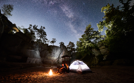 Young couple hikers man and woman having a rest beside camp, bonfire and glowing tourist tent at night. On background beautiful night starry sky full of stars and Milky way, mountain rocks, treesの写真素材