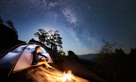 Hiker man resting on rocky mountain top beside camp, campfire and tourist tent at summer night, enjoying magical view of night sky full of stars and Milky way. On background starry sky and treesの写真素材
