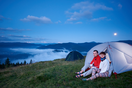 Guy and girl are sitting in a tent at dawn with a view of the beautiful landscape of the morning mountains in a light haze under blue sky with a bright moon and rare cloud.の写真素材