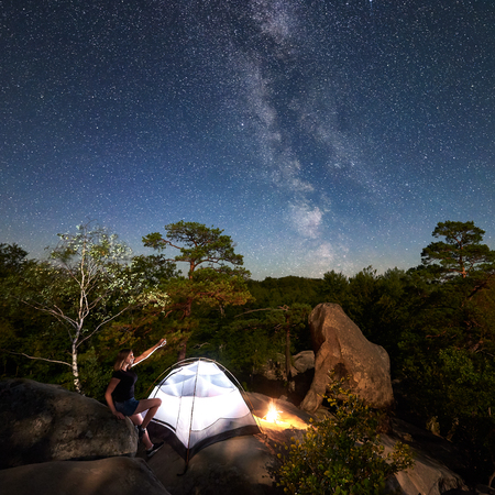 Young woman hiker having a rest on rocky mountain top beside camp, campfire and glowing tourist tent at summer night, pointing at sky full of stars and Milky way. On background boulders and treesの写真素材