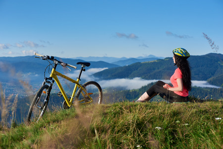 Happy attractive woman biker sitting on the top of mountain near yellow bicycle in the morning, wearing helmet and red red t-shirt. Foggy mountains, forests on the blurred background. Copy spaceの写真素材