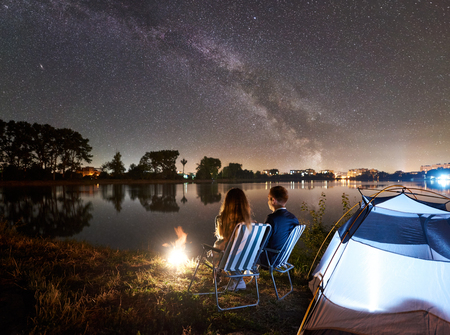 Night camping on lake shore near tent and bonfire. Back view of man and woman tourists sitting on chairs, enjoying incredible view of night sky full of stars and Milky way, city lights on background.の写真素材