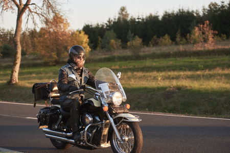 Bearded biker in sunglasses, helmet and black leather clothing riding cruiser motorcycle along empty asphalt country road by beautiful tall trees with golden foliage on sunny autumn evening.の写真素材