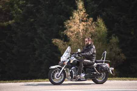 Side view of bearded long-haired motorcyclist in sunglasses and black leather clothing riding cruiser motorbike along narrow asphalt path on sunny autumn day on background of forest.の写真素材