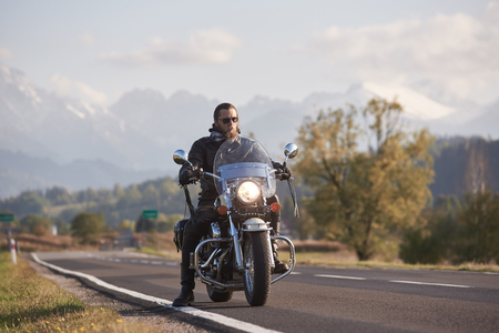 Portrait of handsome bearded biker in black leather jacket on cruiser motorcycle on country roadside on blurred background of green woody hills, distant white mountain peaks.の写真素材