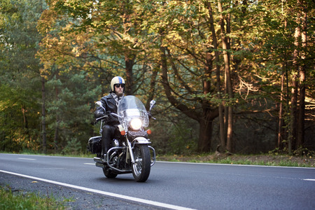 Handsome bearded biker in sunglasses, helmet and black leather clothing riding cruiser motorcycle along asphalt road winding among tall green trees on sunny summer evening.の写真素材