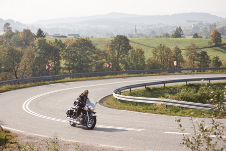 Bearded motorcyclist in helmet, sunglasses and black leather clothing riding bike along sharp turn of empty road on bright summer day on misty background of rural landscape and distant green hills.の写真素材