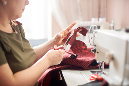 Female hand using scissors for cutting off textile piece. Close up side view of woman, engaging in sewing process at workshop. Blurred background. Handicraft, hobby, self-employment, business concept.の写真素材