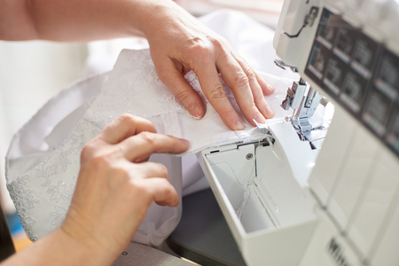 Woman hands working on overlock machine stitching wedding dress at workplace. Seamstress hands holding lace textile for dress production on professional equipment. Blurred background. Close up view.の写真素材