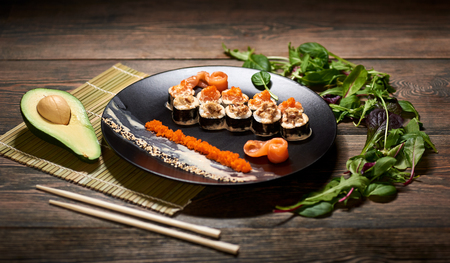 Delicious sushi rolls with caviar and sesame on black plate served on the wooden table, decorated with avocado, chopsticks and sprouts.の写真素材