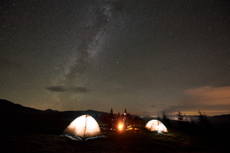 Group of four tourists, men and woman having a rest at night camping by burning bonfire at tourist illuminated tents under night sky with bright sparkling stars and Milky Way constellationの写真素材