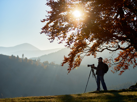 Man tourist photographer with backpack, tripod and professional camera standing under large tree with golden leaves on woody foggy mountains landscape and bright sunset in blue sky background.の写真素材