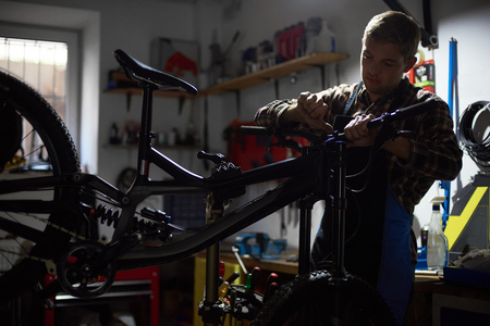 Male mechanic working in bicycle repair shop, serviceman repairing modern bike using special instrument, wearing protective workwear and glovesの写真素材
