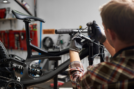Cropped shot back view of professional mechanic working in bicycle repair shop, man repairing modern bike using special tool, wearing protective workwear and glovesの写真素材