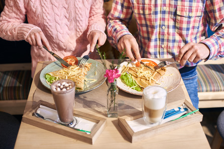 Cropped view of young couple, friends sitting together in cafe at small table enjoying big pancake sandwich with vegetables. Coffee beverage and vase with flower on blurred foreground. High angle viewの写真素材