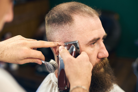 Side view portrait of adult dark-haired bearded man in modern barbershop during haircutting process with electric razor. Professional tools and accessories in barbers handsの写真素材