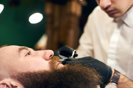Cutting mustache with sharp scissors. Barber in black hygienic gloves concentrate in scrupulous work process. Mustache styling and cutting. Advertising for barber shop. Side view. Blurred background.の写真素材