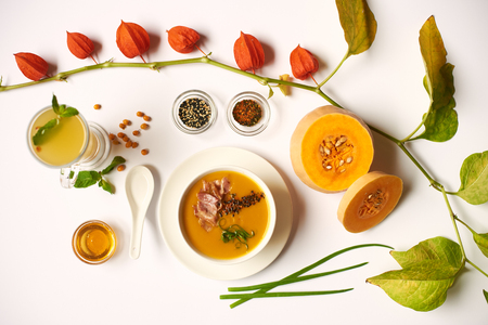 Flat lay of orange cream soup with meat and tea with mint prepared for guest in oriental restaurant. Pumpkin and physalis lying near meal. Above view of healthy food with decor on white tablecloth.の写真素材