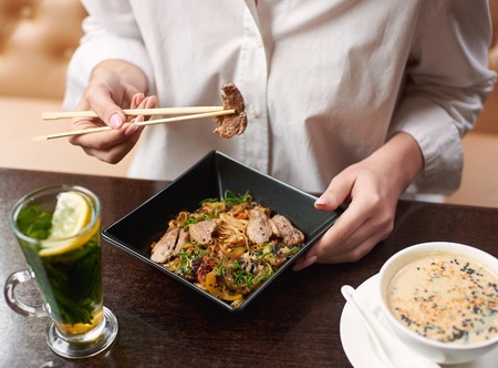Woman ordering oriental style meal in restaurant. Client enjoying lunch including soup with seeds, warm salad with meat and lemon mint tea. Close up hands with manicure holding food sticks.の写真素材