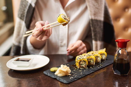 Woman covered in blanket relaxing and eating dinner in restaurant. Girl with manicure enjoying Japanese dish with soya sauce using food sticks. Oriental style lunch serving with wasabi and ginger.の写真素材