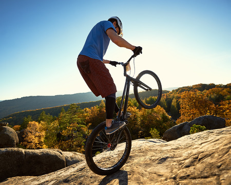 Athlete cyclist standing on back wheel on trial bicycle. Sportsman biker making acrobatic trick on the edge of big boulder on the top of mountain at sunset. Concept of extreme sport active lifestyleの写真素材