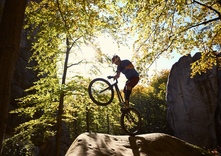 Professional sportsman cyclist riding on back wheel on trial bike, man rider balancing on the edge of big boulder in the forest on sunny day. Concept of extreme sport active lifestyleの写真素材