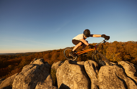 Active man cyclist balancing on trial bicycle on top of big boulder, rider making acrobatic trick on summer evening, blue sky and forest on background. Concept of extreme sportの写真素材