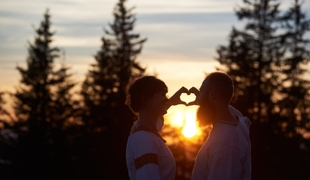Silhouettes of happy couple in love in the evening. Bearded man and smiling woman gesturing heart shaped hands, looking at each. Beautiful sunset, cloudy sky and forest on blurred backgroundの写真素材