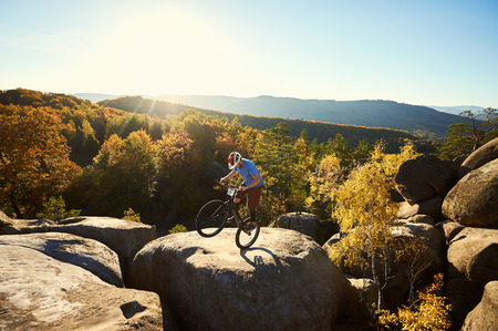 Professional cyclist riding on back wheel on trial bike, sportsman making acrobatic stunt on the edge of big boulder on the top of mountain at sunset. Concept of extreme sport active lifestyleの写真素材
