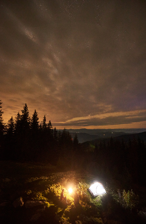 Vertical shot, top view of night summer camping in the mountains. Young couple hikers sitting together beside campfire and illuminated tourist tent under beautiful night cloudy sky with stars.の写真素材