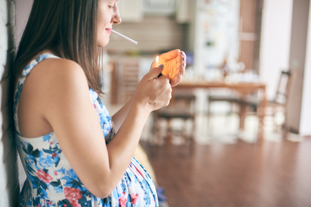 Cropped view of pregnant woman in dress standing in kitchen and keeping in hands cigarette and lighter. Future mother smoking and leading unhealthy lifestyle. Concept of threat and mortality.の写真素材