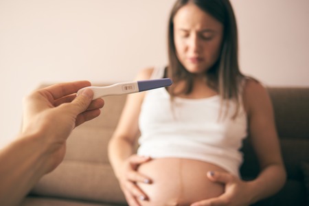 Selective focus of pregnancy test in male hand and disappointed young woman sitting on sofa and keeping belly with hands at background. Concept of despair and unwanted child.の写真素材
