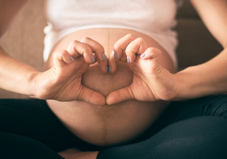 Selective focus of female hands showing sign of heart near big belly. Pregnant woman in white shirt and black pants expecting little baby and enjoying motherhood. Concept of parental love.の写真素材