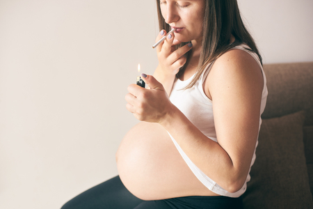 Close up view of lighter and cigarette in hands of pregnant woman. Young future mother in white shirt smoking during pregnancy period. Concept of threat and harmfulness.の写真素材