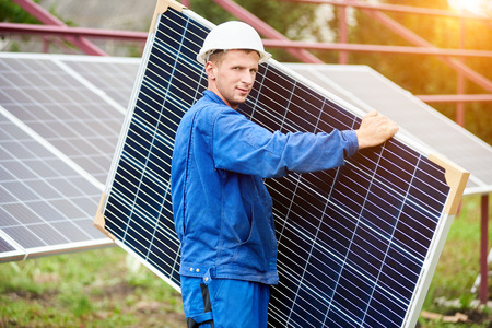 Young worker in blue uniform and protective helmet carrying big shiny solar photo voltaic panel to almost finished exterior metal platform on sunny summer warm day. Renewable ecological green energy.の写真素材