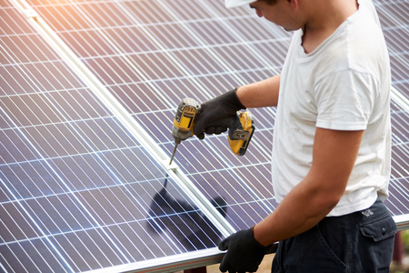 Back view of young technician in helmet connecting solar photo voltaic panel to metal platform using electrical screwdriver on shiny surface background. Stand-alone solar panel system installation.の写真素材