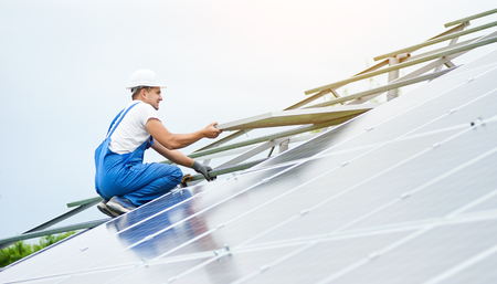 Installing of solar photo voltaic panel system. Professional worker in hard-hat and blue overall lifting the solar module on metal platform. Alternative energy and professional construction concept.の写真素材