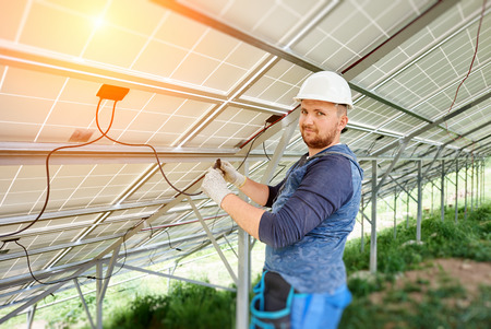 Installing and wiring of stand-alone solar photo voltaic panel system. Close-up of young electrician in hard-hat connecting electrical cables inside the solar modules. Alternative energy concept.の写真素材