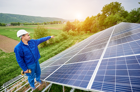 Construction worker with screwdriver standing on metal frame of photo voltaic solar system pointing on shiny surface. Alternative energy, ecology protection and cheap electricity production concept.の写真素材