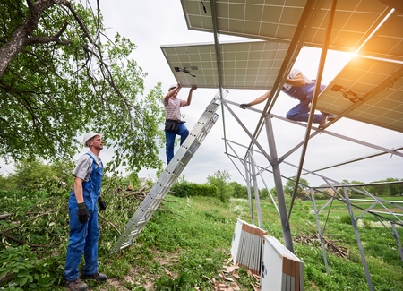 Installing of stand-alone solar photo voltaic panel system. Three technicians lifting big solar module on high metal platform. Investment in alternative energy and professional construction concept.の写真素材