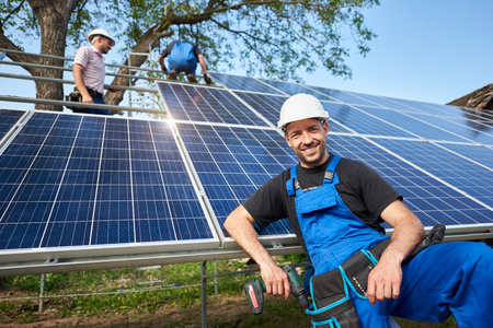 Portrait of smiling technician with electrical screwdriver in front of unfinished high exterior solar panel photo voltaic system with team of workers on high platform.の写真素材