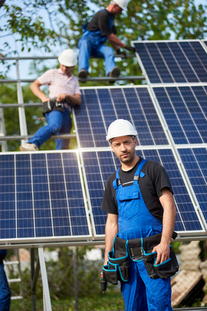 Portrait of engineer technician with electrical screwdriver standing in front of unfinished high exterior solar panel photo voltaic system blue shiny surface with team of workers on high platform.の写真素材