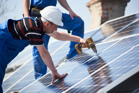 Male engineers installing stand-alone solar photovoltaic panel system using screwdriver. Electricians mounting blue solar module on roof of modern house. Alternative energy ecological concept.の写真素材