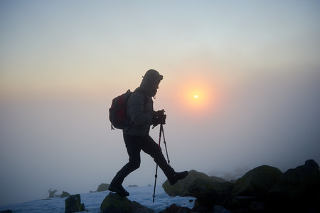 Silhouette of tourist hiker man with backpack and trekking sticks climbing on rocky mountain peak on background of foggy cloudy misty blue sky and raising bright orange sun at dawn.の写真素材