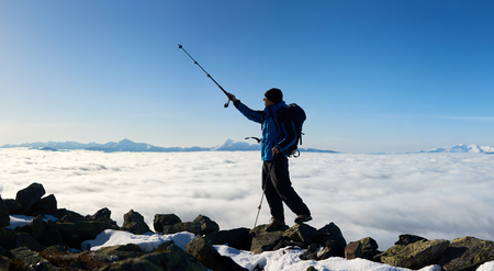 Tourist hiker with backpack standing on huge rocks on the top of mountain pointing with trekking pole at blue sky on background of foggy valley filled with white clouds and distant mountain tops.の写真素材