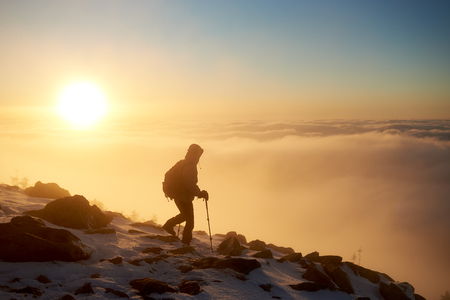 Tourist hiker with backpack and trekking sticks hiking on rocky snowy mountain steep slope on background of foggy valley filled with white puffy clouds, raising sun and blue sky at sunrise.の写真素材